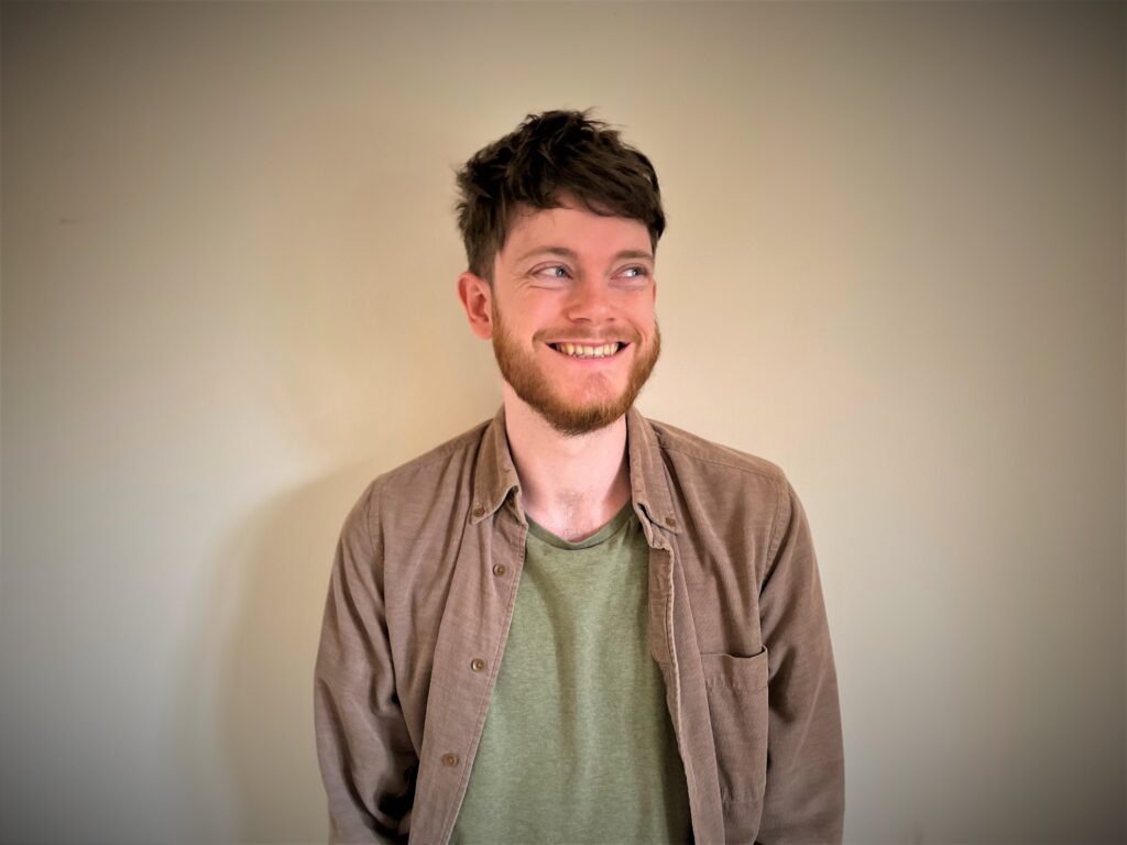 Joe Clarke, a white man with tousled brown hair stands in front of a cream-coloured wall. He's wearing a green t-shirt and a light brown corduroy shirt. He has a big smile on his face and is looking to his left.